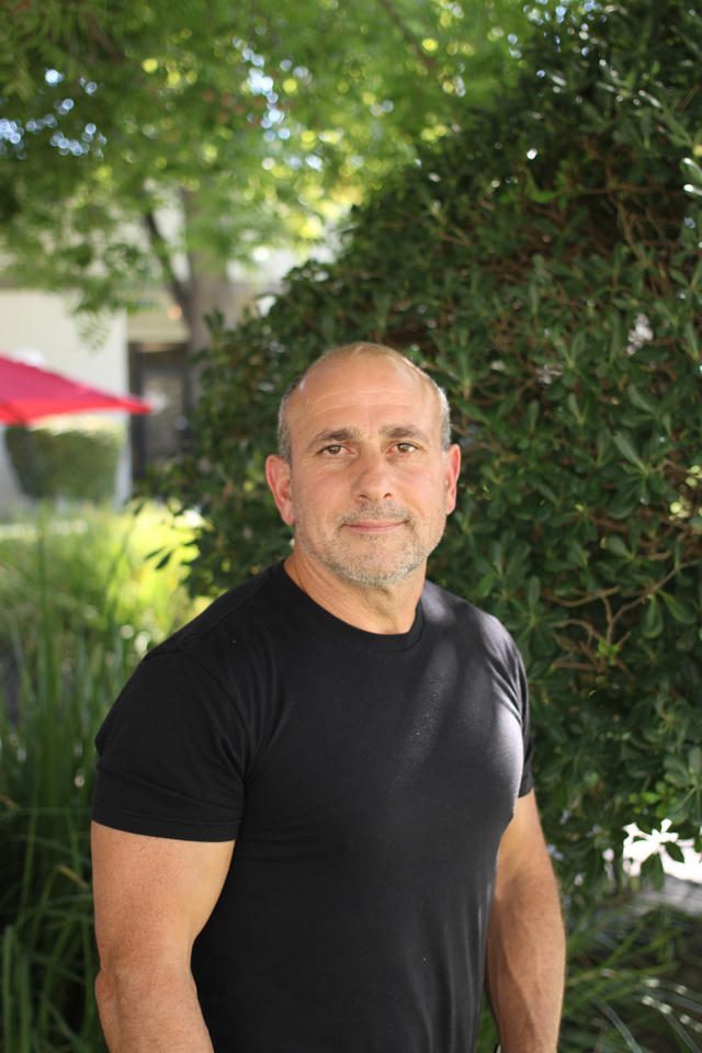 A man in a black t-shirt stands outdoors with greenery and a red umbrella in the background.